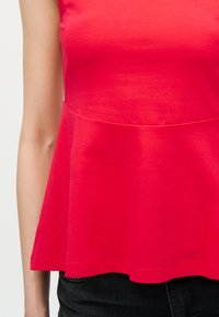 Αμάνικο κόκκινο τοπ πεπλούμ συνδυασμένο με μαύρο παντελόνι, που δείχνει τον κορμό και το αριστερό χέρι σε απλό φόντο.