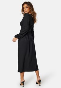 Zwarte jurk met lange mouwen van textuurstof, met een taille die is cinched en een vloeiende rok. Draagt een paar open-taille zwarte hakken, zichtbaar zijsplit.