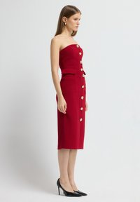 Rode straploze midi-jurk met gouden knoopaccenten en zijzakken. Aansluitende lijfje, rechte rok, textuurstof. Zwarte hoge hakken.