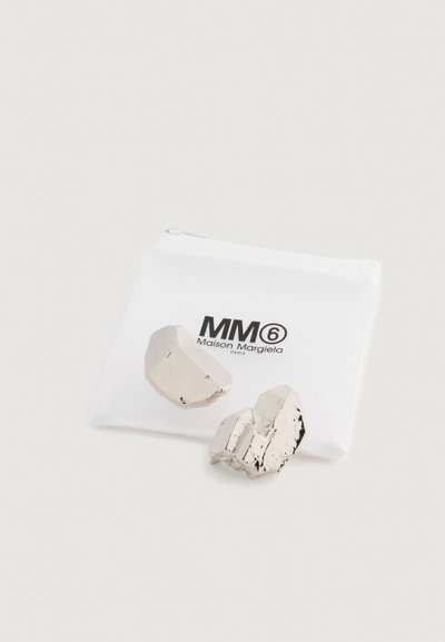 Kovové strieborné šperky s nepravidelnými tvarmi, vystavené vedľa bieleho púzdra s logom MM6 Maison Margiela.