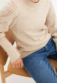 Person i en beige stickad tröja med texturerade puffärmar och blå jeans som sitter på en trästol.