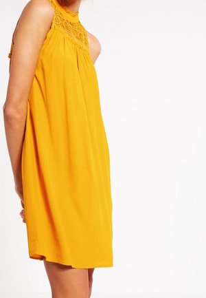 Bezrekawnikowa żółta sukienka z koronkowym zdobieniem wokół dekoltu, noszona przez osobę stojącą na jednolitym białym tle.