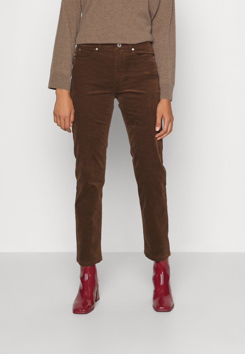 Personne portant un pantalon en velours marron, un haut à manches longues beige et des bottines rouges brillantes, debout devant un fond clair uni.