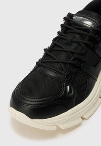 Svart sneaker med en glatt læroverflate og mesh-stoff. Har runde lisser, hvit gummisåle og reflekterende detaljer på tungen.