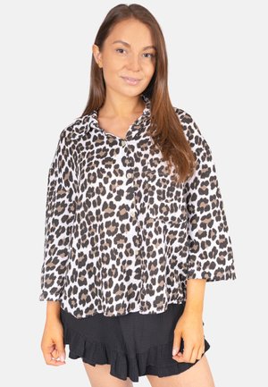 Jeune femme portant une chemise à imprimé léopard boutonnée et un short noir à volants, debout devant un fond clair uni.