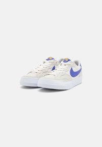 Adidași Nike cu un superior crem, accente albastre, talpă albă și design cu șireturi, realizati din materiale texturate și cu un guler căptușit.