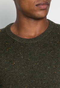 Pull en vert foncé avec une texture côtelée, col rond et petits éclats multicolores dans le tissu. Zone du cou et du menton visible.