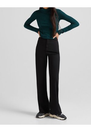 Un col roulé vert côtelé associé à un pantalon large noir taille haute. Le modèle porte des baskets noires ; textures lisses et design ajusté visibles.