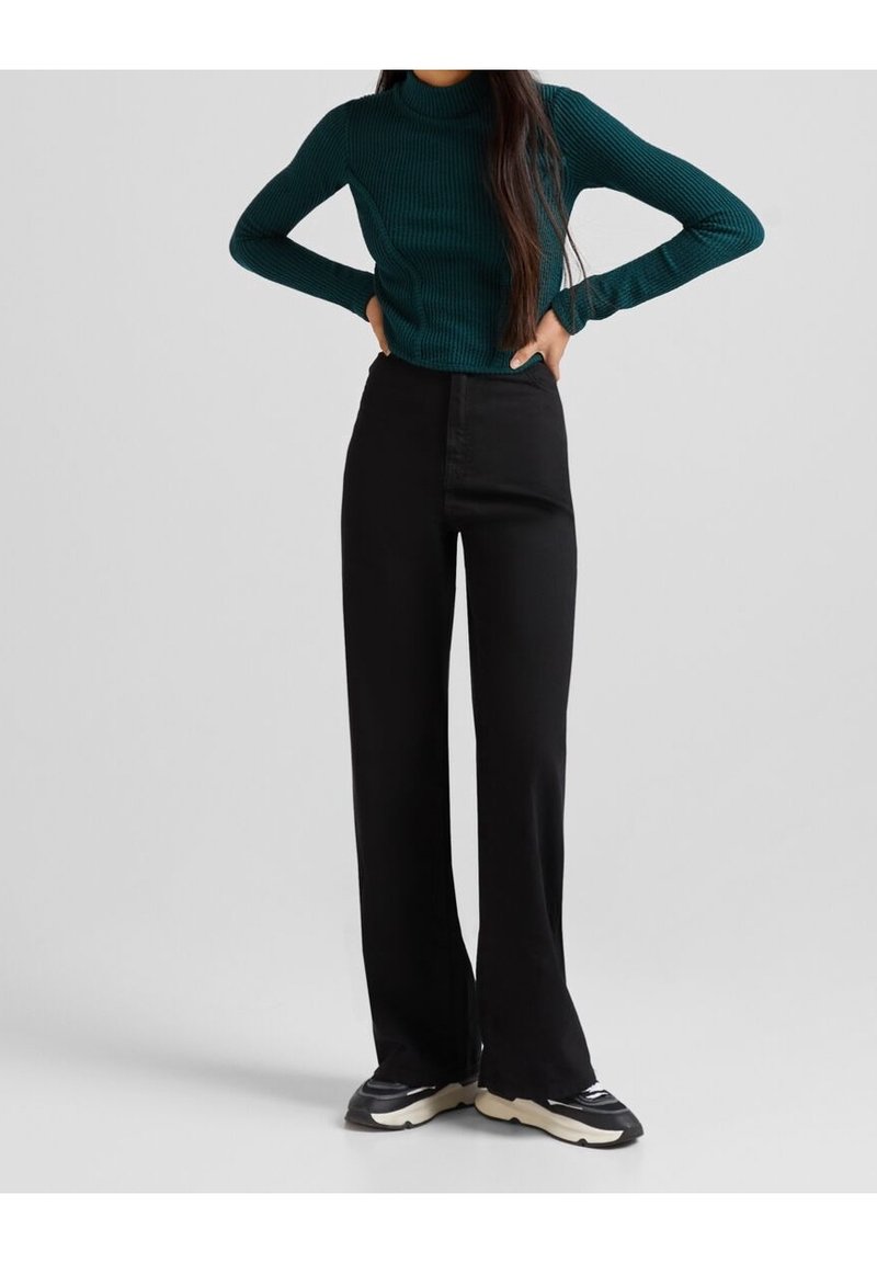 Cuello de tortuga verde acanalado combinado con pantalones de pierna ancha negros de cintura alta. La modelo lleva zapatillas negras; texturas suaves y diseño ajustado visibles.