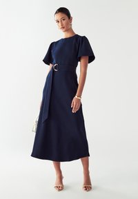 Námorníckomodrá midi šaty s krátkymi puff rukávmi, opásaným pásom s zlatým prstenom a prúdiacou sukňou. Nosí sa s telovými podpätkami a bielou kabelkou.