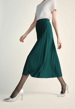 Zielona plisowana spódnica zestawiona z białym t-shirtem i srebrnymi szpilkami. Materiał wydaje się zwiewny o gładkiej fakturze.