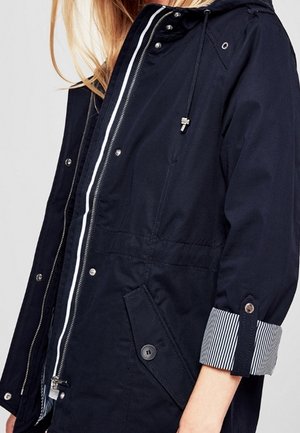 Frau trägt eine marineblaue Kapuzenjacke mit aufgesetzten Pattentaschen mit Knöpfen und hochgekrempelten Ärmeln, die ein gestreiftes Futter zeigen.