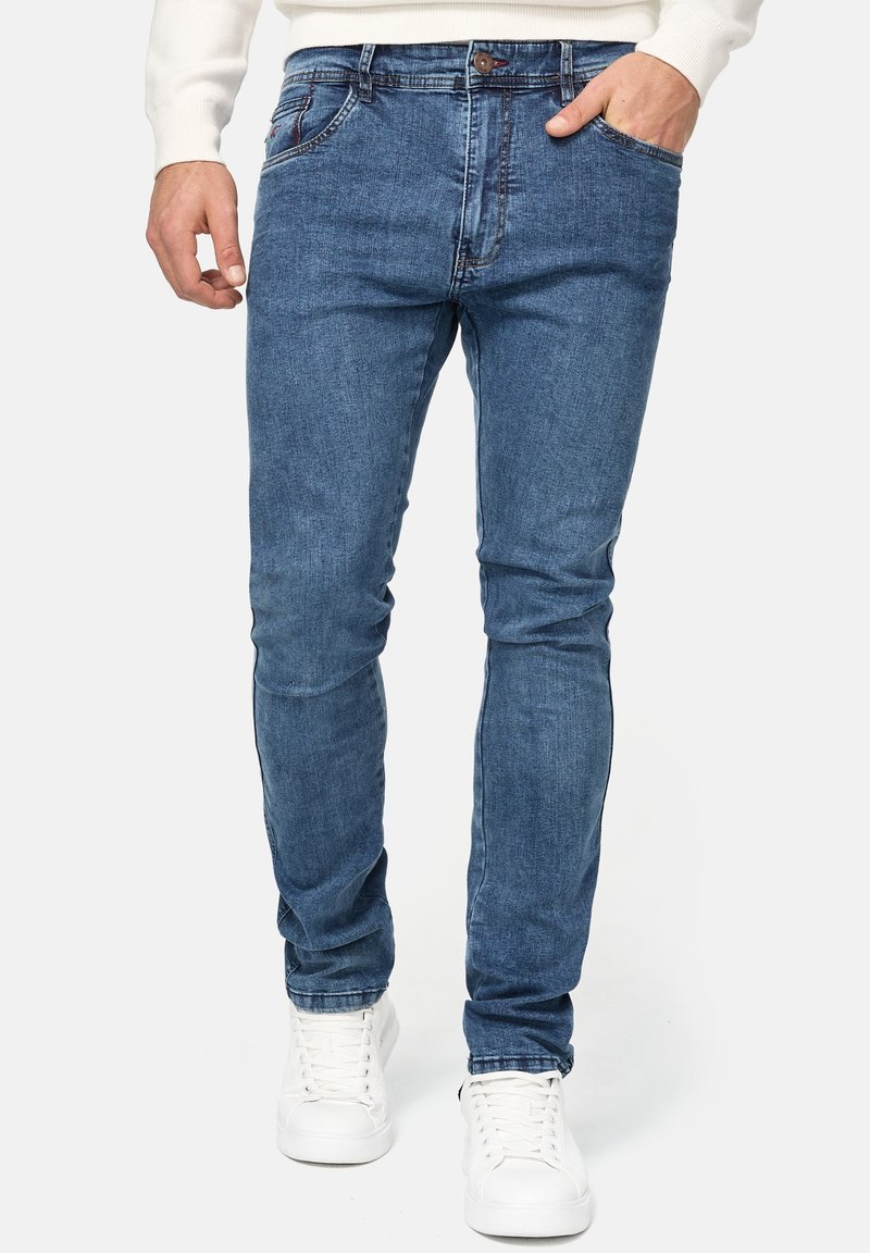 Jean en denim bleu slim-fit pour homme avec une texture lisse, cinq poches, une fermeture à bouton sur le devant, et des baskets blanches.