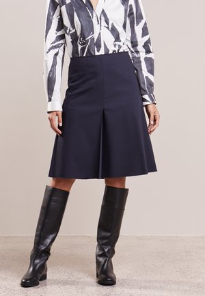 Vrouw die een zwart-wit patroon lang shirt met lange mouwen draagt, een marineblauwe knielange rok en zwarte kniehoge leren laarzen, staand op een gestructureerde vloer.