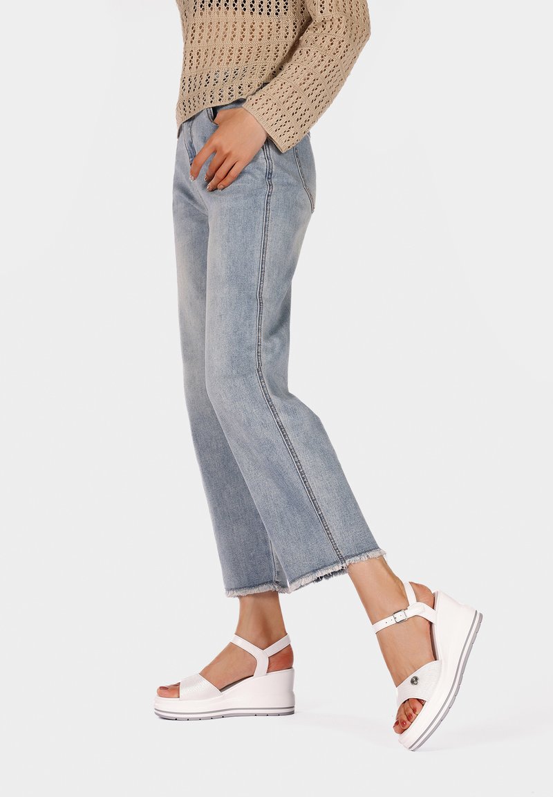 Sandali con zeppa bianchi con cinturini texturizzati, un accento di fibbia e una suola piatta, abbinati a jeans azzurri con orlo sfrangiato e a un top in maglia beige.