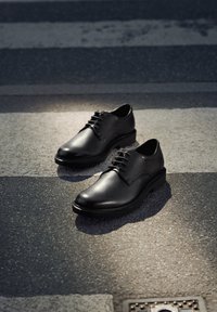 Par de zapatos de vestir de cuero negro pulido colocados sobre una carretera asfaltada con líneas blancas de paso de peatones.