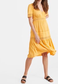 Mujer con un vestido amarillo hasta la rodilla con detalles de encaje, de pie con una mano en la cadera, usando sandalias planas negras sobre un fondo blanco.