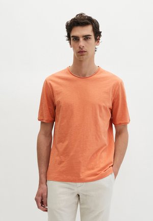 Jeune homme aux cheveux bouclés portant un t-shirt orange uni à manches courtes et un pantalon beige clair, debout devant un fond blanc.