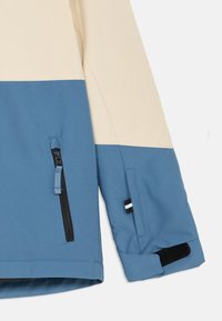 Blaue und cremefarbene Jacke aus glattem Material, mit einer Reißverschlusstasche und Klettverschluss-Manschetten. Zweifarbiger Blockdesign mit kontrastierenden Nähten.