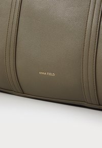Olivově zelená texturovaná taška z umělé kůže s vertikálním prošíváním a kovovým zlatým logem 'ANNA FIELD'.