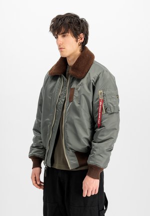 Young man with tousled dark hair wearing a grey bomber jacket with brown fur collar and a red zipper tag on the sleeve.