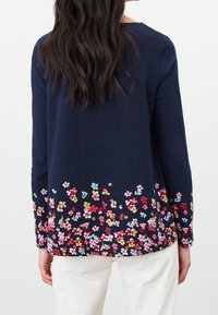 Marinblå långärmad topp med en blommönstring i rosa, rött, gult och blått vid fållen. Mjuk textur och avslappnad passform. Vita byxor synliga.