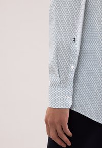 Weißes Hemd mit hellblauem Muster, langen Ärmeln, Knopfmanschetten und einem kleinen schwarzen Stickdetail am Ärmel. Das Muster zeigt kleine geometrische Formen.