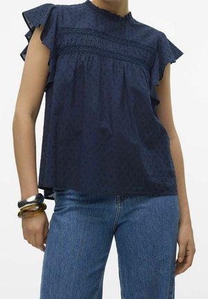 Person iført en mørkeblå bluse med flæser på ærmerne og strukturerede detaljer, kombineret med blå jeans og to chunky armbånd på det ene håndled.