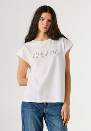 Junge Frau mit langen dunklen Haaren trägt weißes ärmelloses Pepe Jeans T-Shirt und blaue Jeans, steht vor einem einfarbigen hellen Hintergrund.