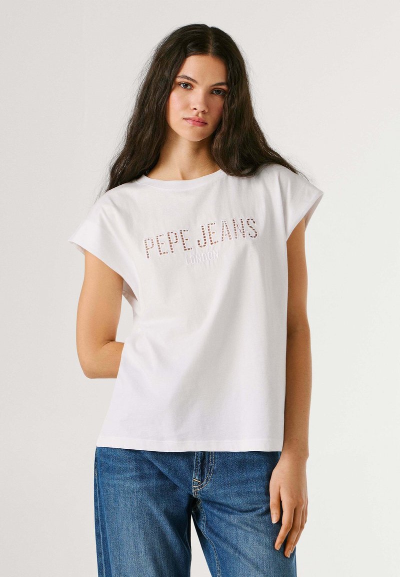 Młoda kobieta o długich ciemnych włosach nosi białą, bezrękawnikową koszulkę Pepe Jeans i niebieskie dżinsy, stojąc na tle jednolitego jasnego tła.