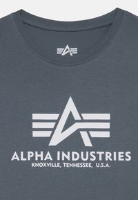 Tamsiai pilka medvilninė marškinėliai su balta logotipu, turinčiu kampuotą dizainą, įskaitant horizontalius raštus, ir užrašu "ALPHA INDUSTRIES."
