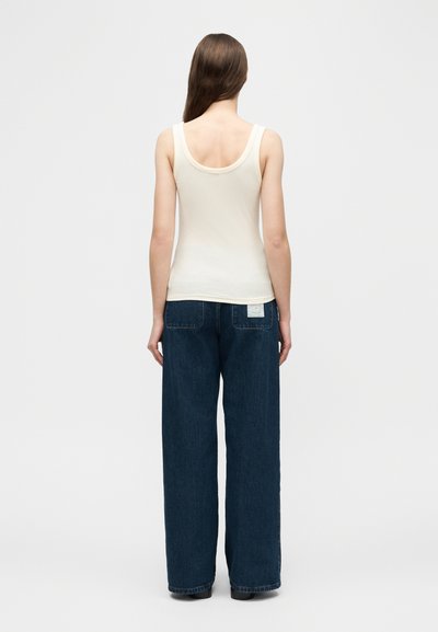 Femme aux longs cheveux bruns portant un haut sans manches crème et un jean large bleu foncé, debout de dos sur fond blanc.