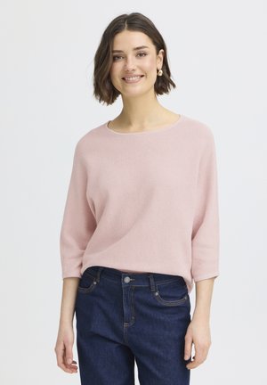 Mladá žena s tmavými krátkymi vlasmi sa usmieva, má na sebe svetloružový top s troma štvrťovými rukávmi a tmavomodré džínsy, stojí pred jednofarebným pozadím.