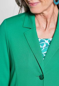 Grüner Blazer mit glatter Textur, Reverskragen und einem einzigen Knopfverschluss. Darunter ein gemustertes Oberteil in Türkis und Weiß.