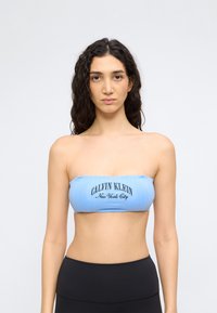 Γυναίκα με μακριά σγουρά μαλλιά φοράει ανοιχτό μπλε τοπ χωρίς τιράντες Calvin Klein και μαύρα ψηλόμεσα εσώρουχα, στέκεται μπροστά σε απλό φωτεινό φόντο.