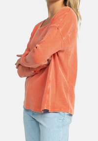 Haut à manches longues orange clair texturé, coupe ample, coutures visibles et tissu côtelé. Associé à un pantalon en denim bleu clair.