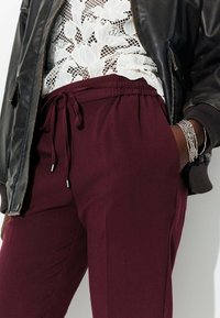Pantalons stretch bordeaux avec une taille élastique et un cordon de serrage, dotés de poches latérales, associés à un haut en dentelle et une veste bomber noire.