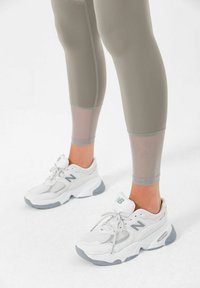 Hvite sportssko med nett- og lærdetaljer, med grå aksenter. Leggings i lys beige med gjennomsiktige paneler ved leggene.