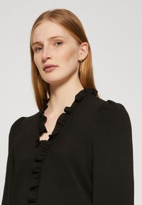 Black top with ruffled neckline, long puff sleeves, and textured fabric. Model wearing gold hoop earrings against a plain background.