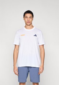 Camiseta blanca de manga corta con texto "TENNIS" en naranja y logo de Adidas en azul oscuro, combinada con pantalones cortos azul claro, mostrando un ajuste relajado.