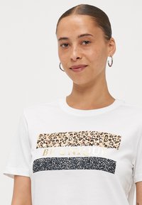 T-shirt en coton blanc avec trois bandes horizontales aux motifs léopard en beige et noir, et le texte "SOIS UNIQUE" en métal doré.