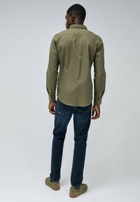 Camisa de manga larga verde olivo con puños de botón, combinada con jeans azul oscuro y zapatos de ante verdes, vista desde atrás.
