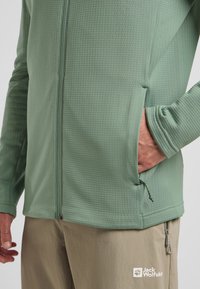 Veste zippée vert clair en tissu texturé, avec des poches latérales, des poignets élastiques et une coupe ajustée. Les logos sur le tissu sont visibles.