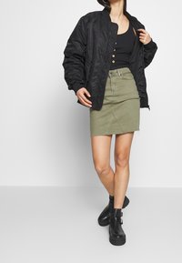 Chaqueta bomber acolchada negra, falda de mezclilla verde oliva con dobladillo deshilachado y botas negras de tobillo con hebillas. El conjunto tiene un diseño casual.