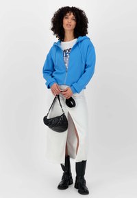 Blauwe zip-up hoodie, witte graphic t-shirt, witte rok met split, zwarte gewatteerde handtas en zwarte knielange laarzen.