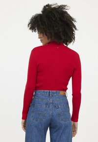 Roter gerippter Rollkragenpullover mit langen Ärmel, kombiniert mit hochgeschnittenen blauen Jeans aus Denim, die über gebogene Nähte an den hinteren Taschen verfügen.