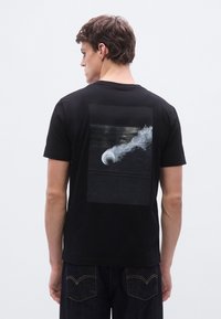 Μαύρο βαμβακερό μπλουζάκι με γραφικό τύπωμα στην πλάτη που απεικονίζει ένα υφασμένο μαύρο ορθογώνιο με ένα κυκλικό αντικείμενο και εφέ καπνού.