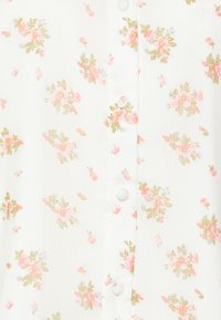 Blusa branca com um padrão floral em rosa e verde. Apresenta botões arredondados ao longo do centro, com um tecido leve e texturizado.