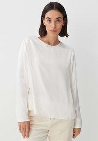 Blusa bianca a maniche lunghe con scollatura rotonda e texture liscia; presenta dettagli di cucitura sottili e una vestibilità comoda.