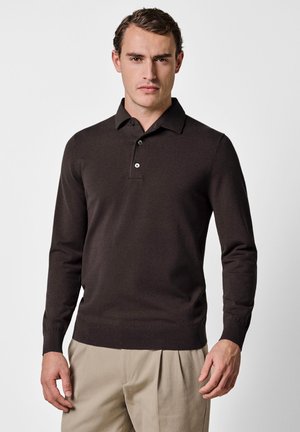 Uomo che indossa una polo a maniche lunghe marrone scuro e pantaloni beige, in piedi contro uno sfondo chiaro e neutro.